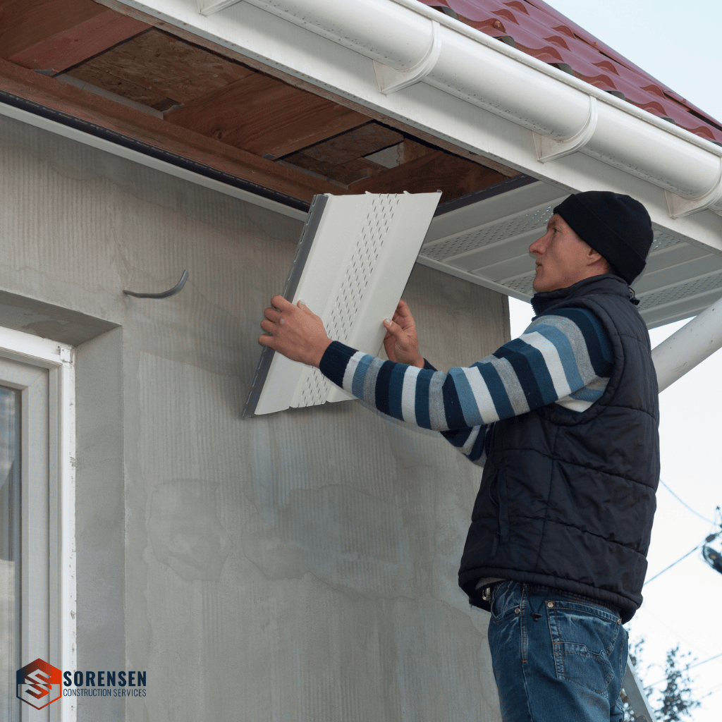 Man in a striped sweater and beanie installing continuous soffit vents under an eave focused on manual work.